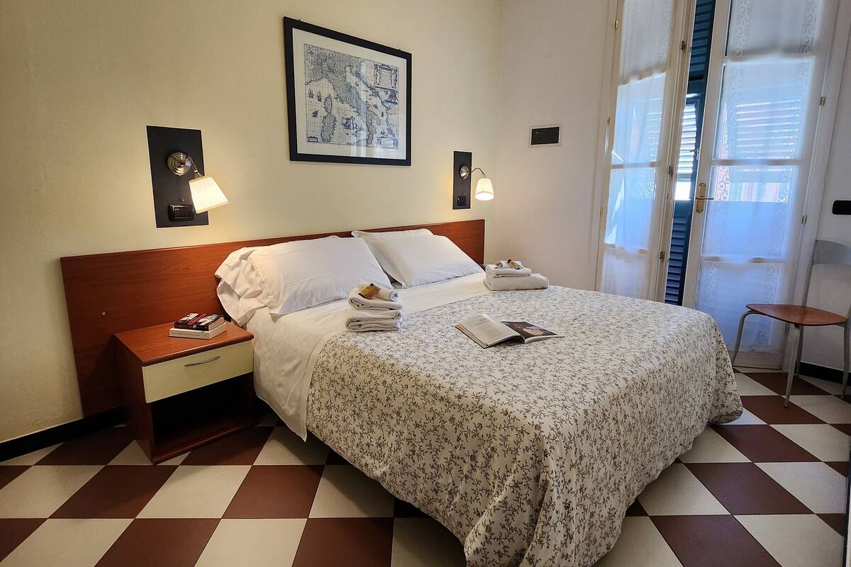 A hotel room with a checkered floor, a bed with a floral bedspread, a nightstand with a lamp, and a framed picture on the wall.