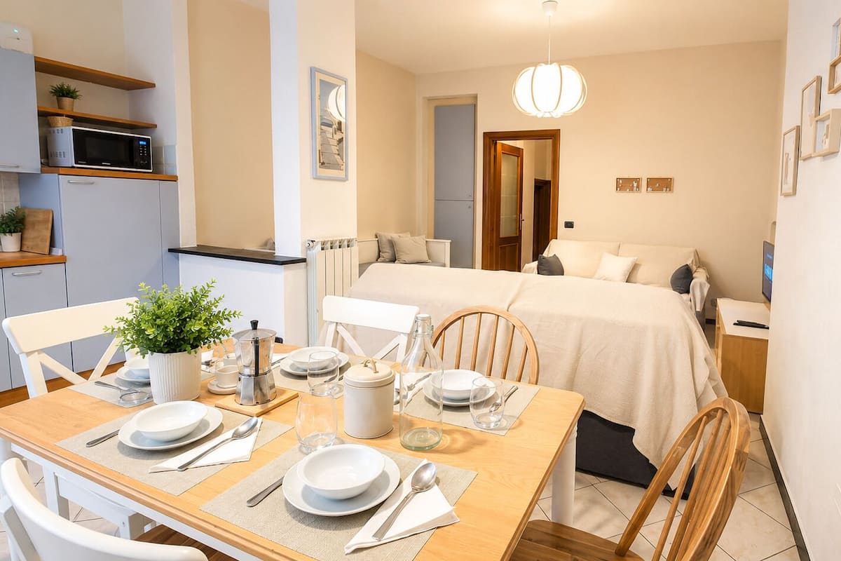 A compact hotel room with a kitchenette, a dining area with a table set for two, and a bed with a white bedspread.
