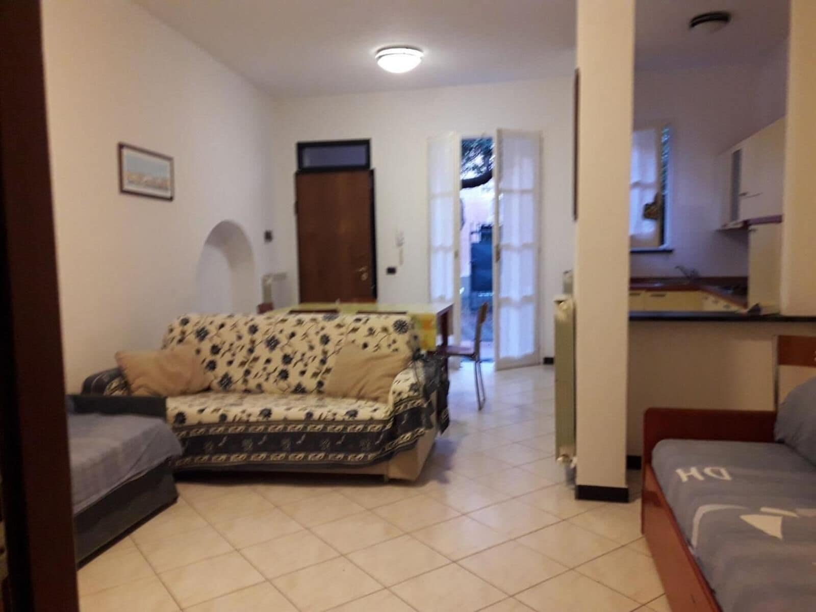 A living room with a sofa, a dining table with chairs, a kitchen area, and a door leading to another room.