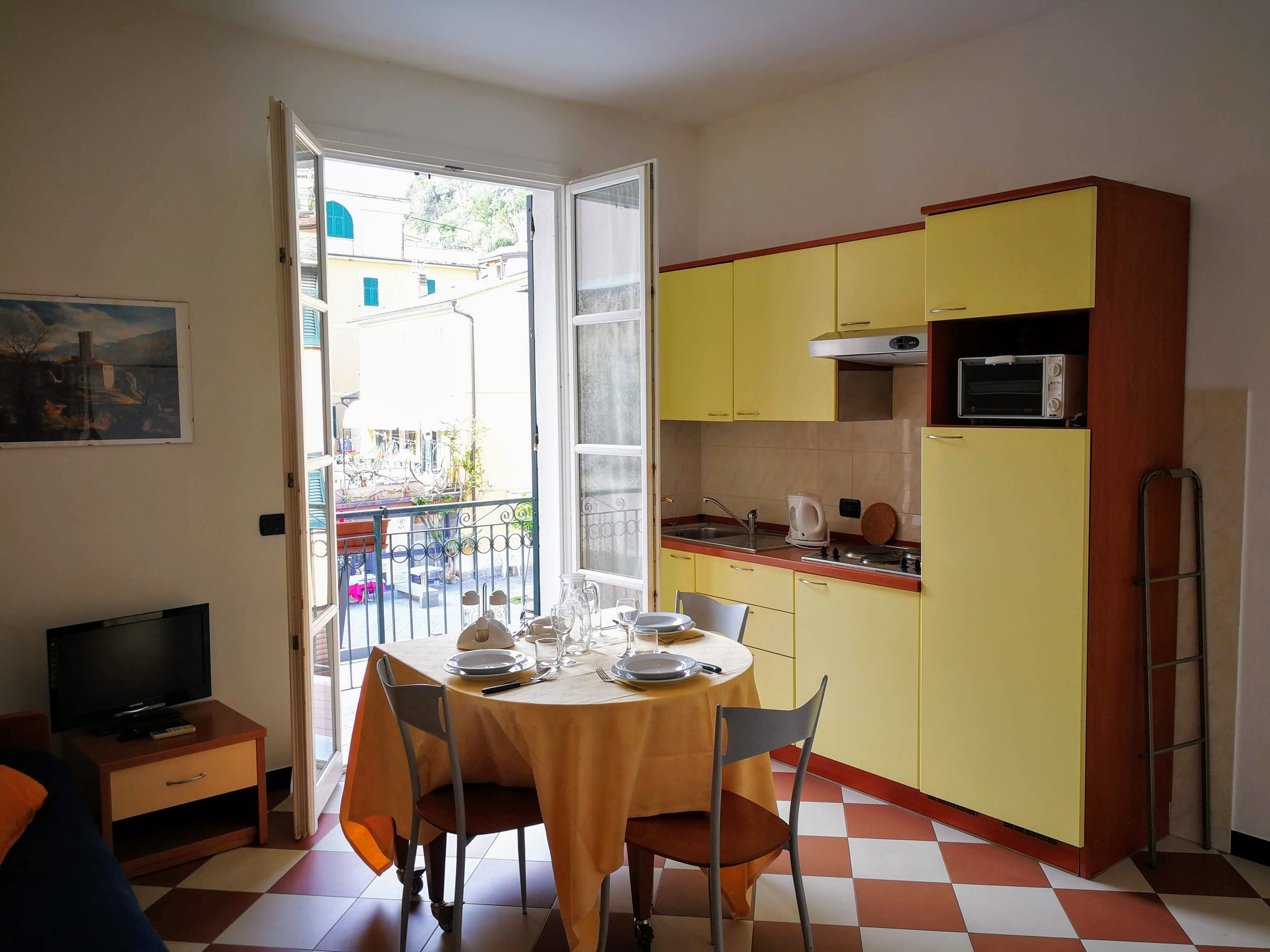 Una cuina senzilla amb armaris grocs, una taula de dinar preparada amb plats i gots, i un televisor sobre una taula petita.