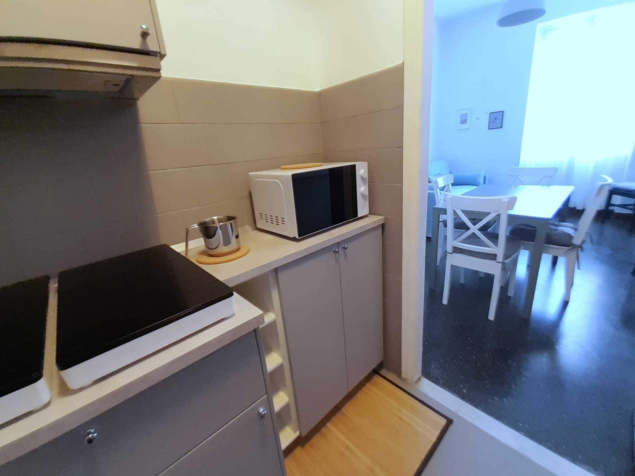 A compact kitchen with a microwave, a kettle, and a stove. There is a dining area with a table and chairs visible through an open door.