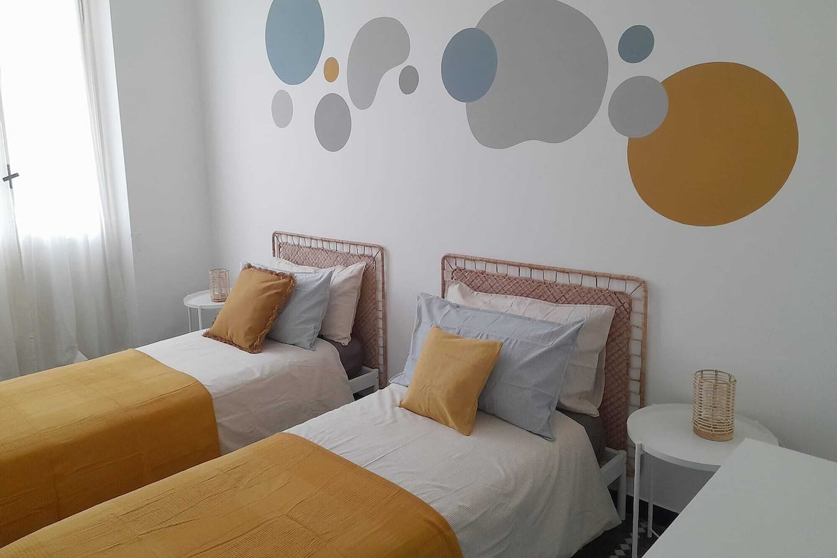 A hotel room with two beds, each with a headboard and pillows, a small round table with a basket, and a wall with abstract art.