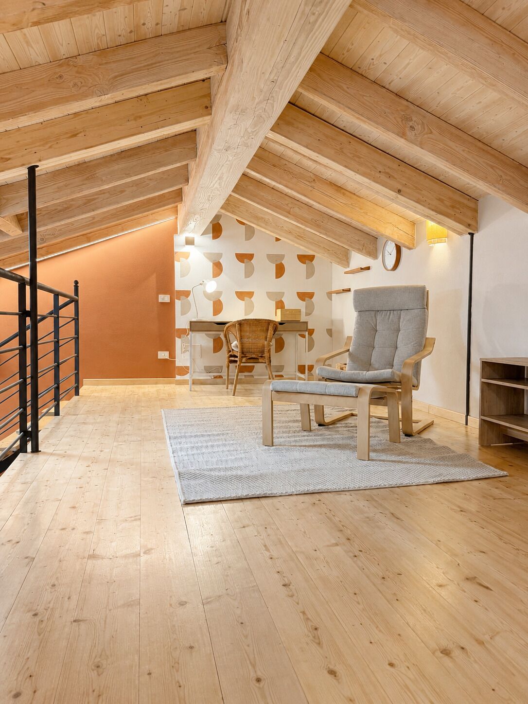 A loft-style room with a high ceiling, wooden beams, a desk with a chair, a fireplace, and a staircase.