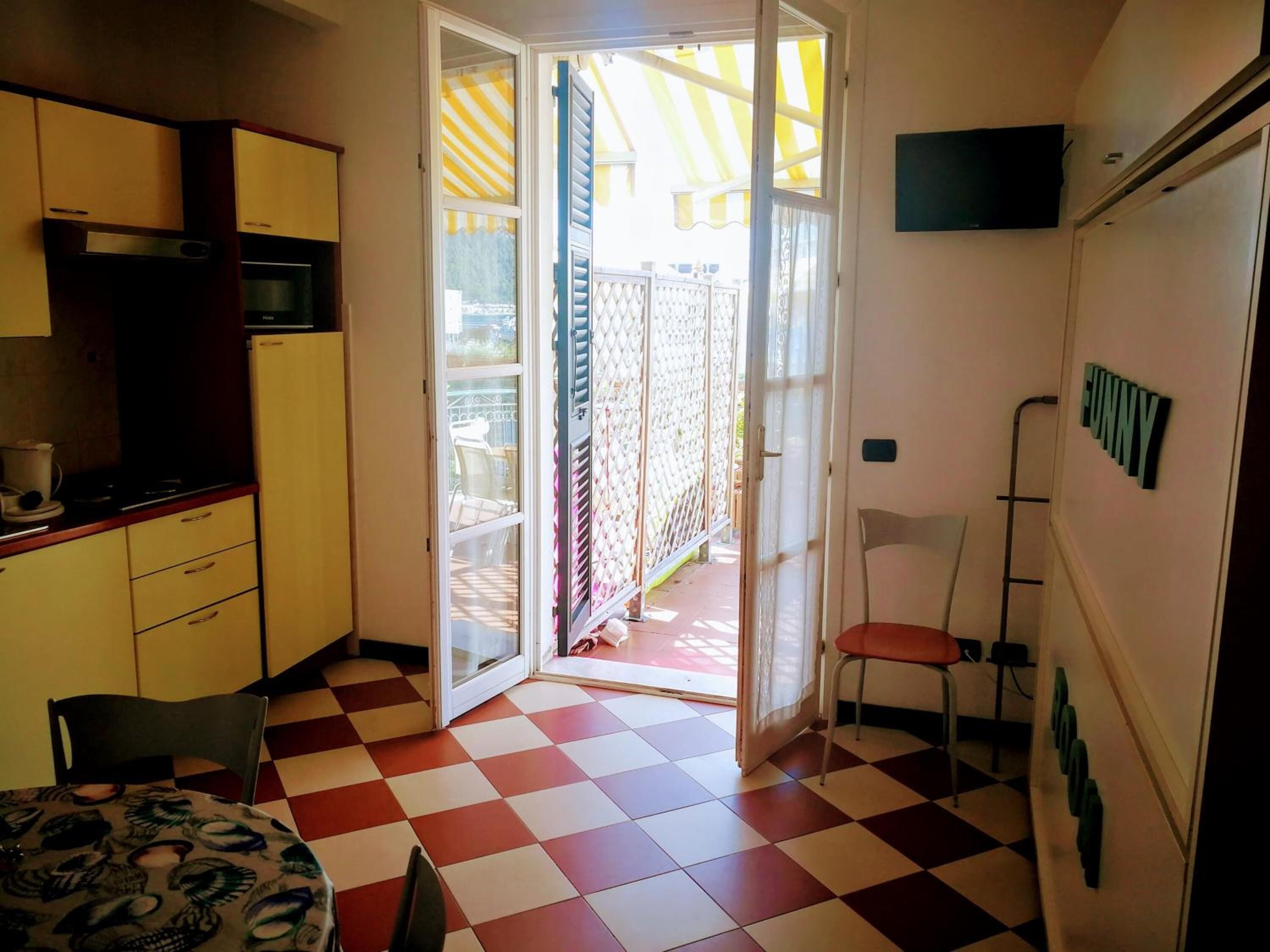 A compact kitchen with a microwave, stove, and a small dining table with chairs. A sliding glass door leads to a balcony with a yellow and white striped awning.
