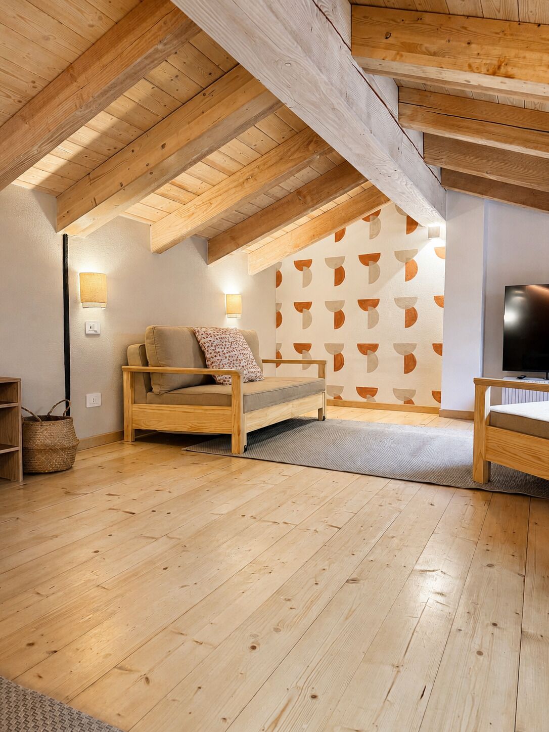 Une chambre à coucher confortable, aménagée dans un grenier avec des poutres apparentes, comprenant un lit, un téléviseur et un mur décoré d’un motif.
