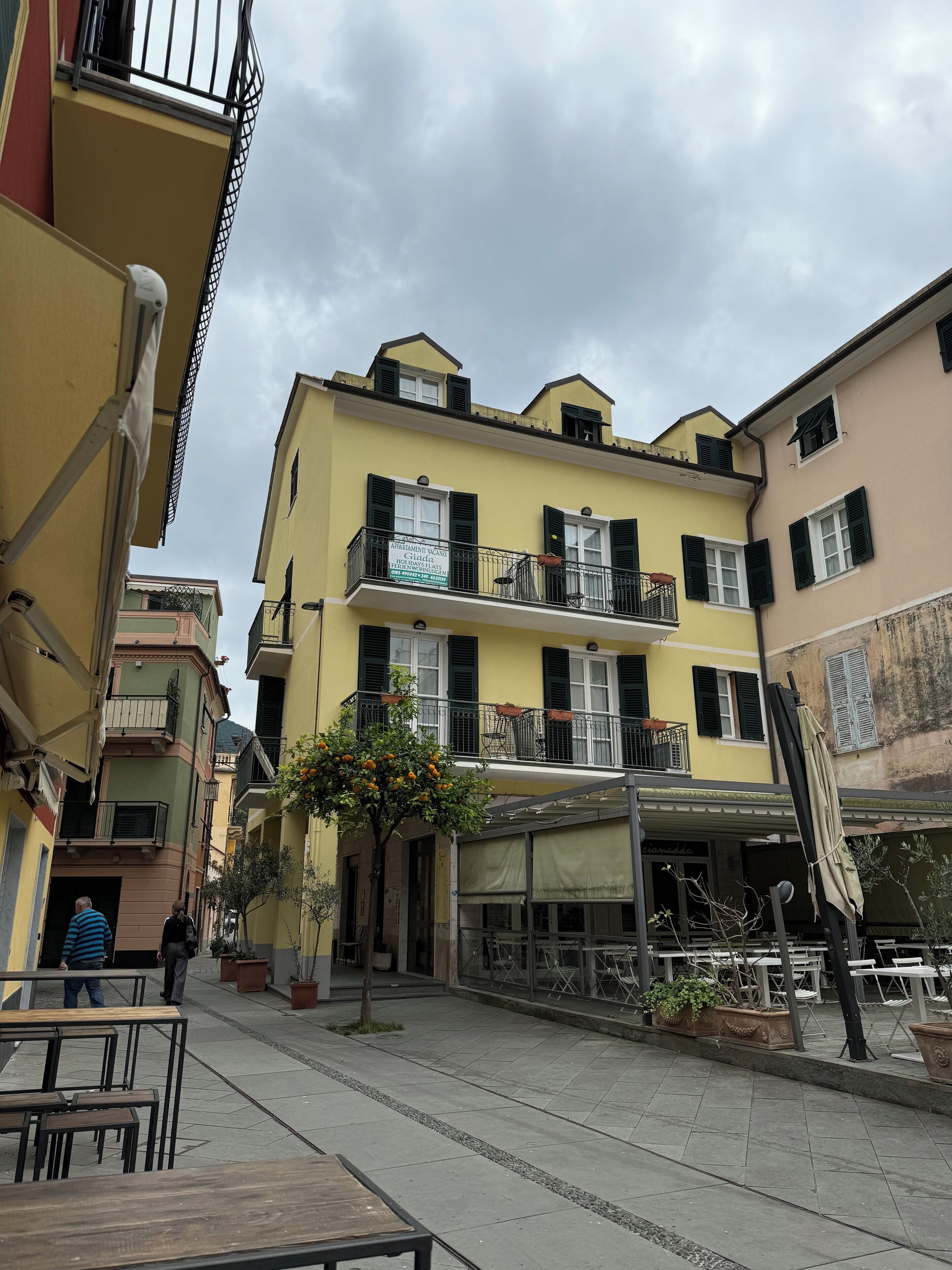 A yellow building with balconies, a tree with orange fruits, and outdoor seating.