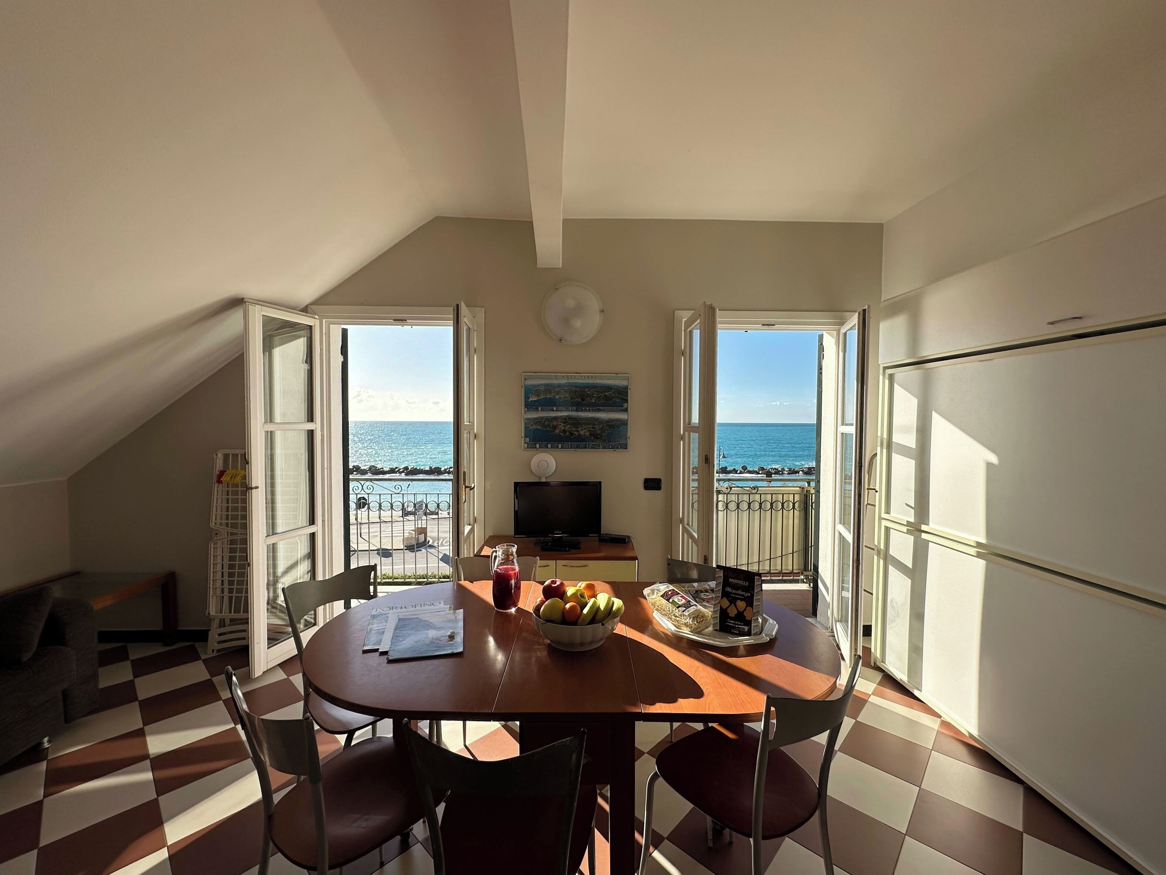 A dining area with a view of the sea, a table with chairs, a TV, and a refrigerator.