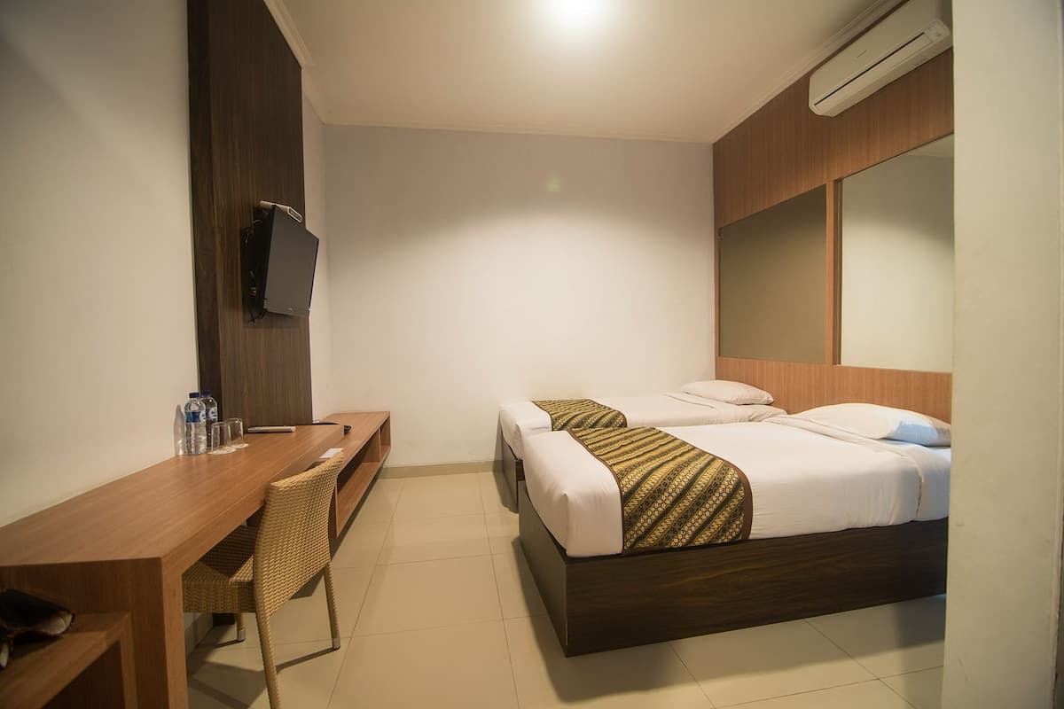 A hotel room with a wooden desk, two beds with patterned bedspreads, a television, and an air conditioning unit.