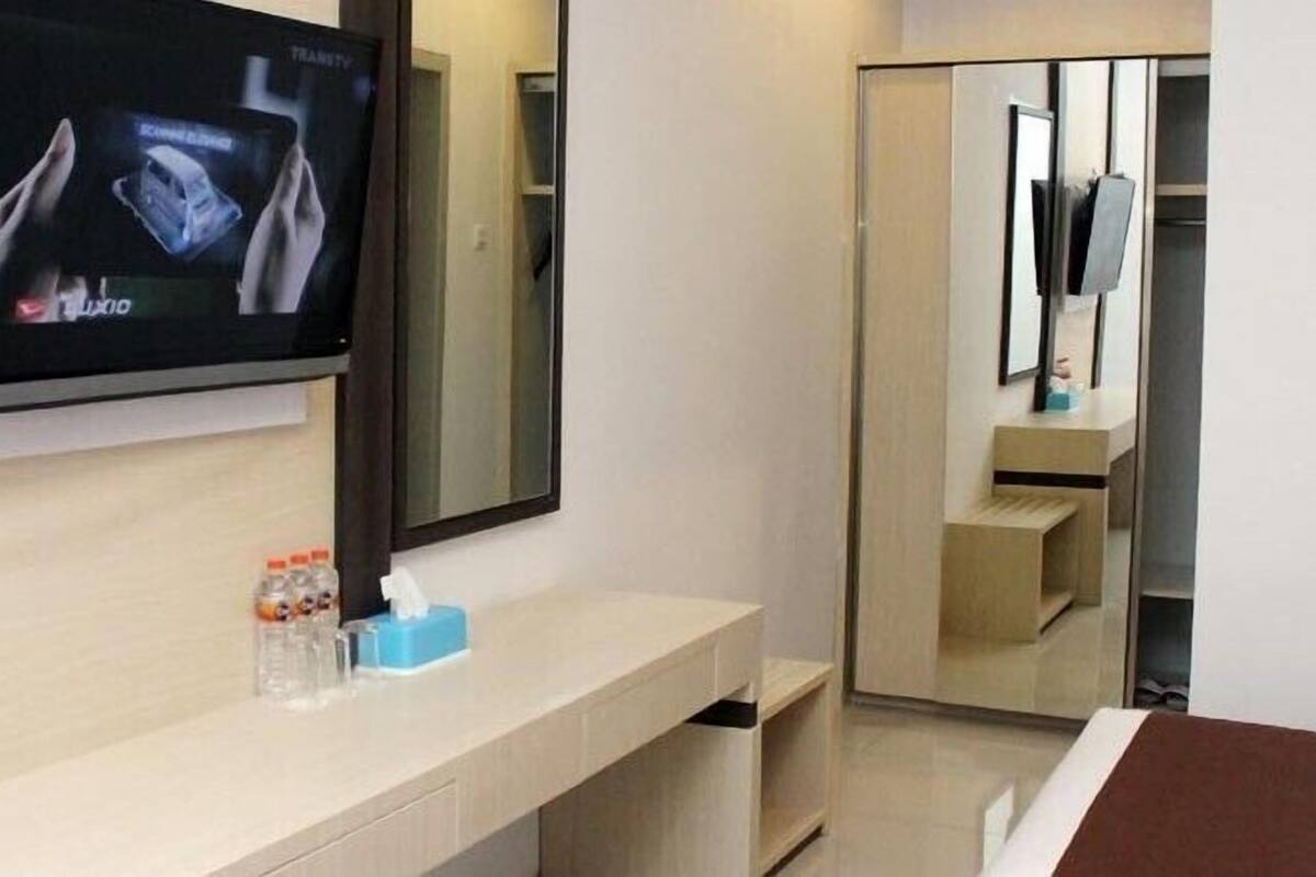 A hotel room with a TV mounted on the wall, a bedside table with a tissue box and water bottles, and a mirror above the table.