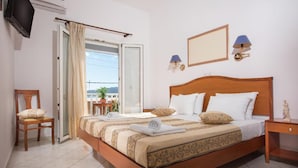 A hotel room with a wooden bed, a chair, a TV, a balcony with curtains, and a wall-mounted lamp. - Meltemi Hotel (Milos)
