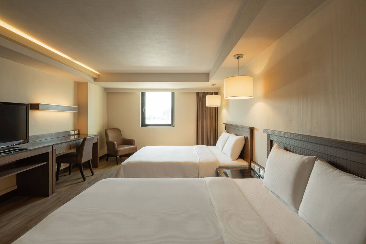 Ein Hotelzimmer mit zwei Betten, einem Schreibtisch mit Fernseher, einem Sessel und einem Fenster mit Vorhängen.
