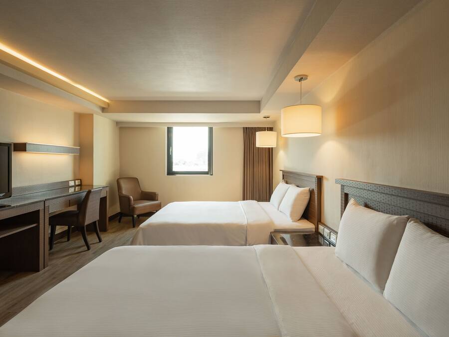 A hotel room with two beds, a desk with a TV, a chair, and a window with curtains.