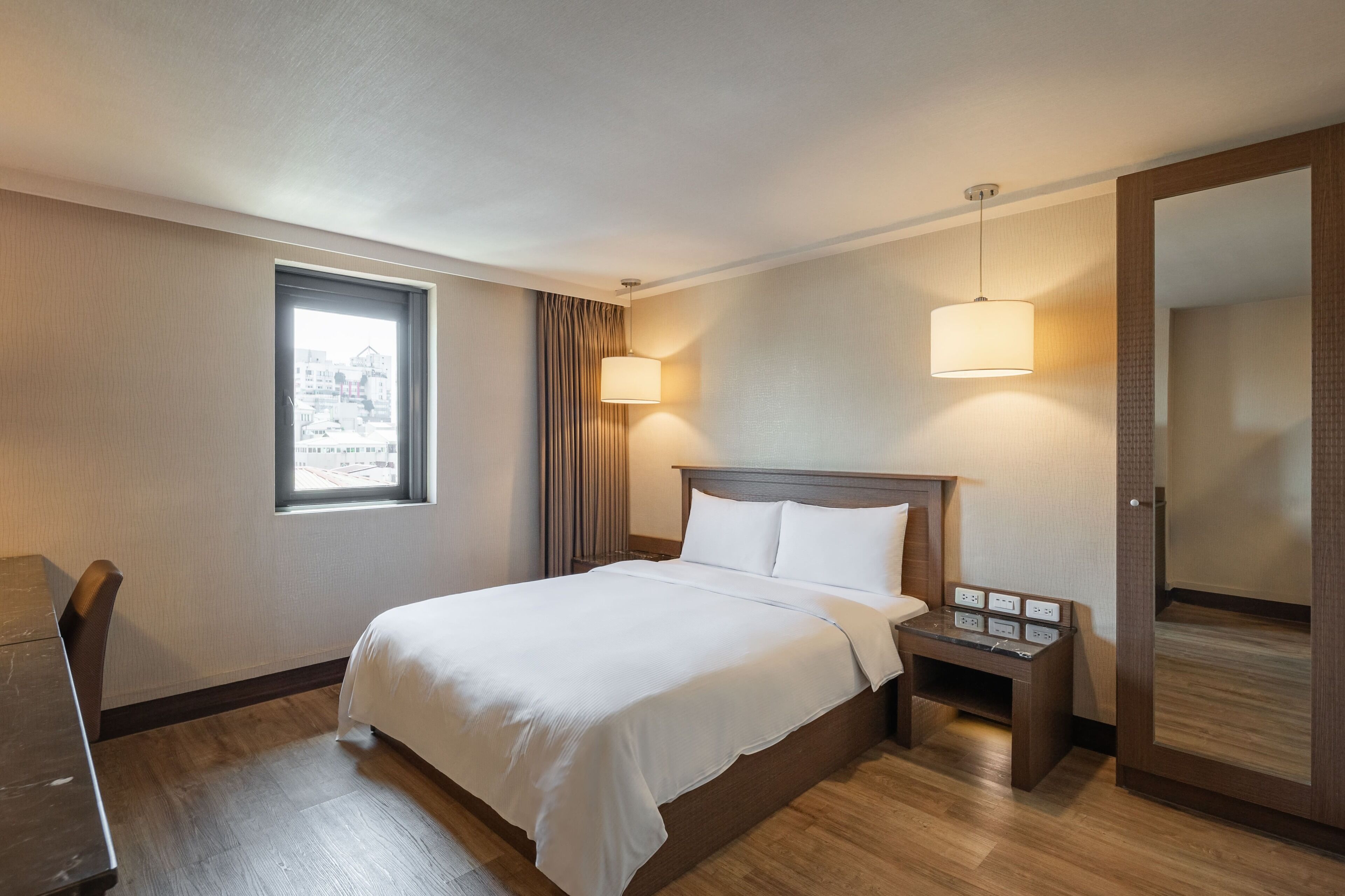 A hotel room with a bed, bedside table, lamp, window, and a mirror.