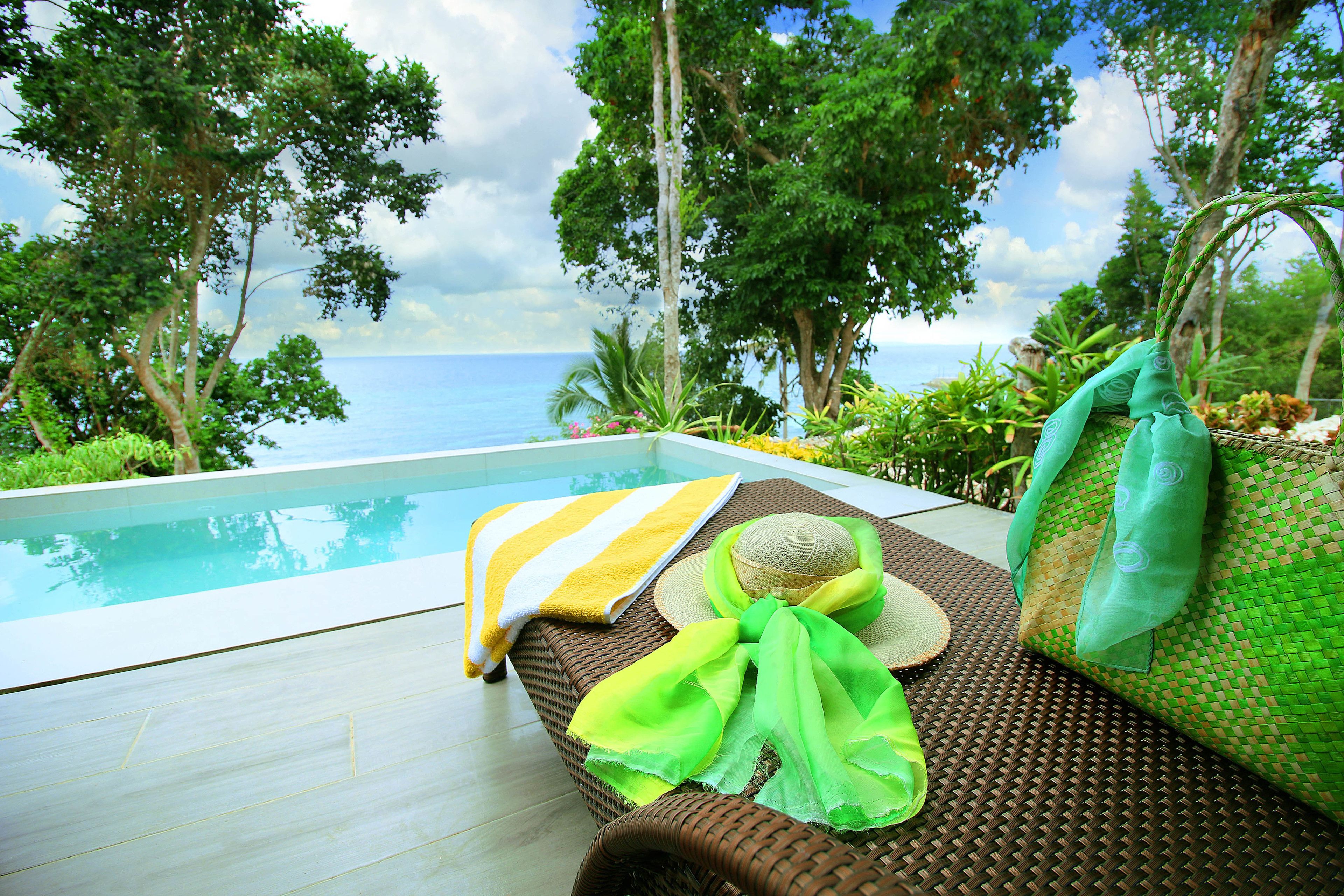 A wicker lounge chair with a yellow and white striped towel and a green cloth, a straw hat, and a woven pillow, overlooking a pool and a tropical landscape with trees and the sea.