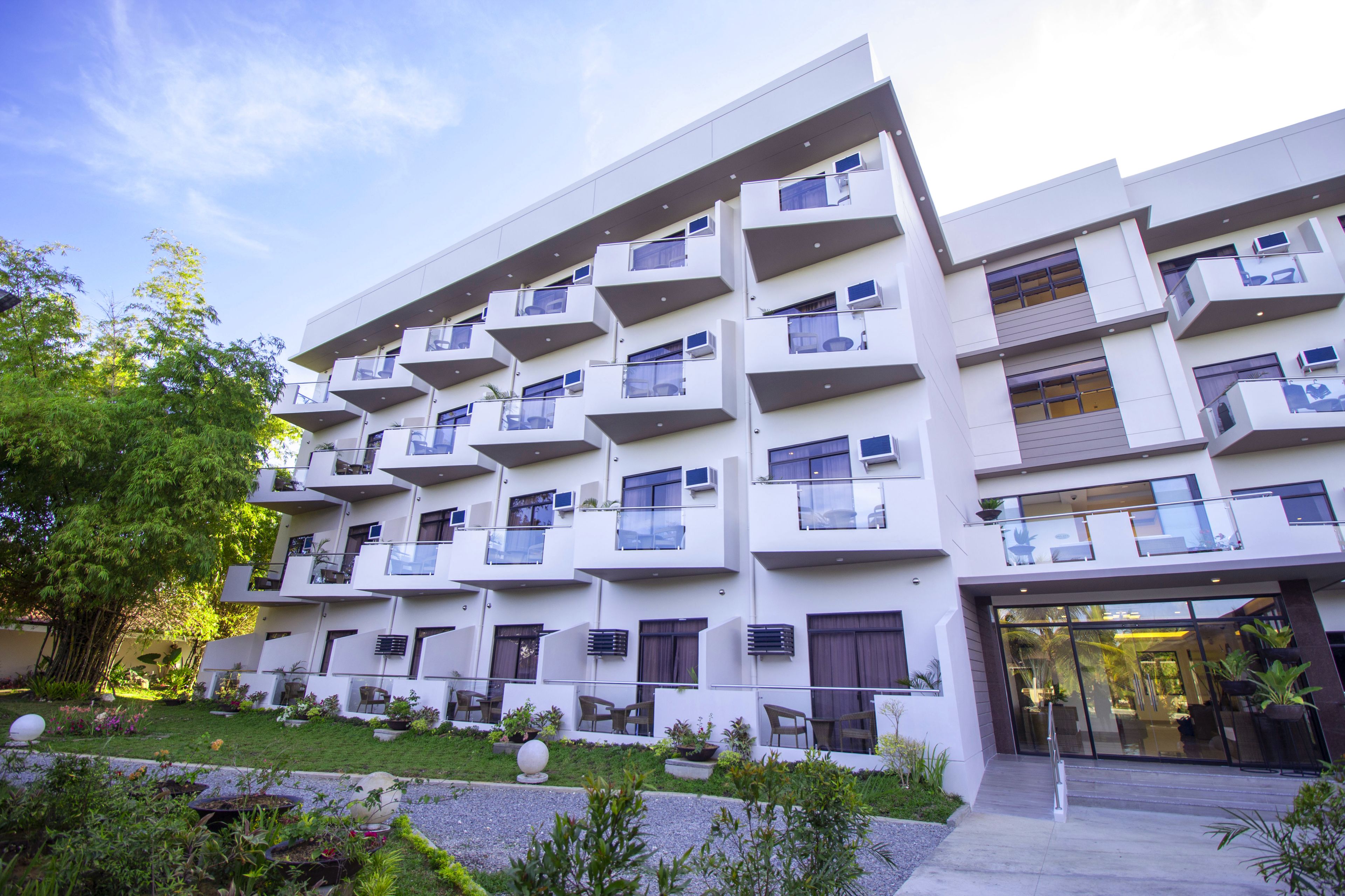 A modern multi-story hotel with balconies, a landscaped garden, and a glass entrance.