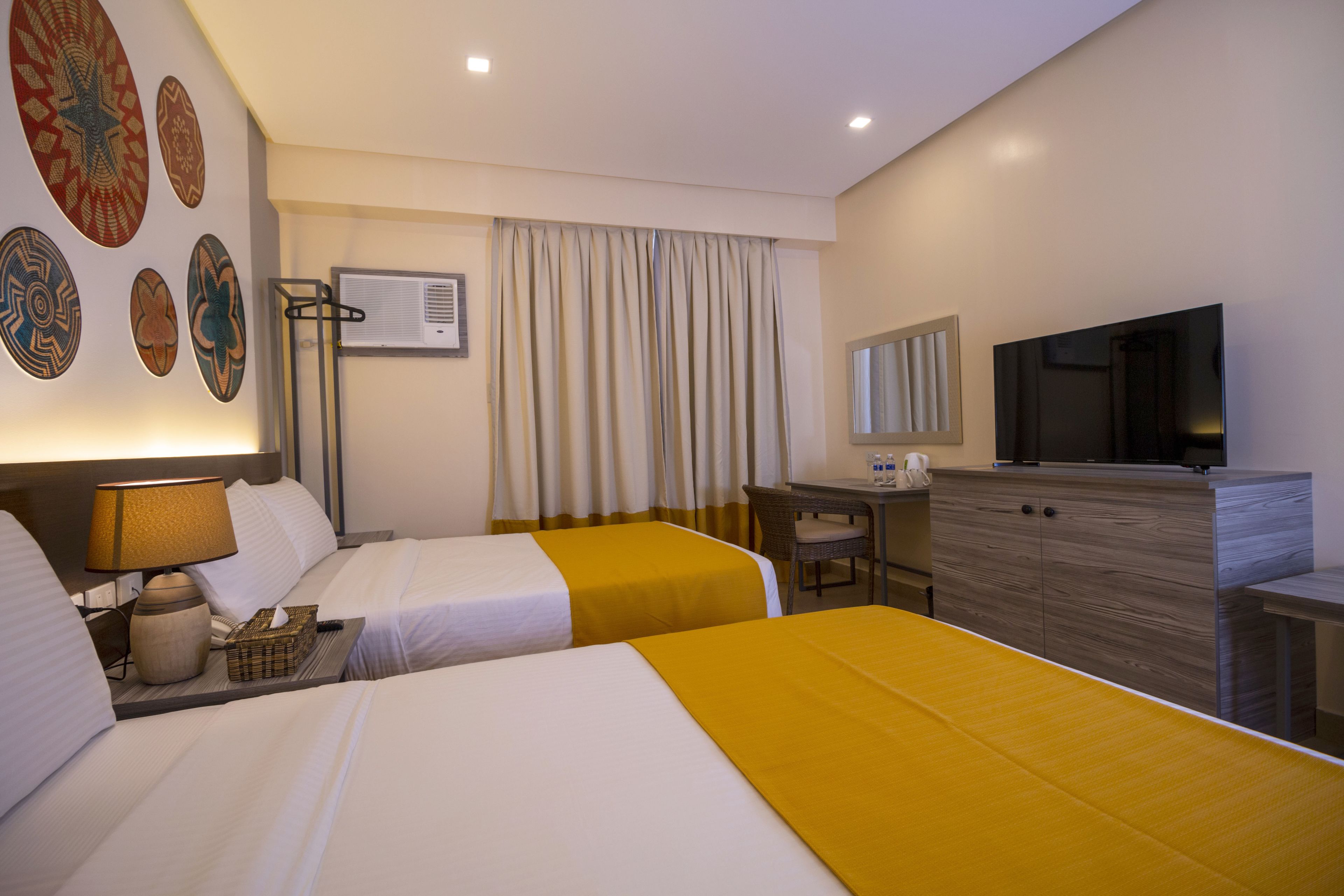 A hotel room with two beds, a desk, a television, and a wall-mounted air conditioner.