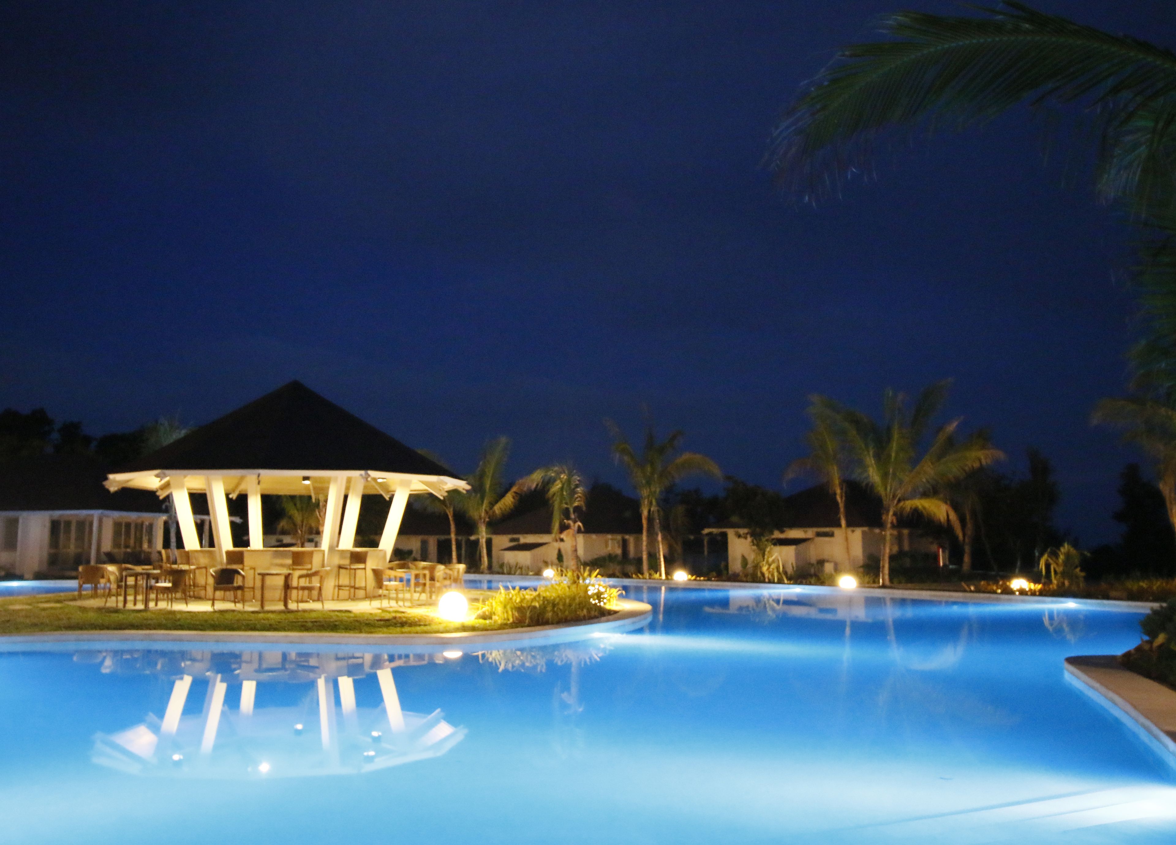 A nighttime pool area with a gazebo, palm trees, and a clear pool reflecting the lights.