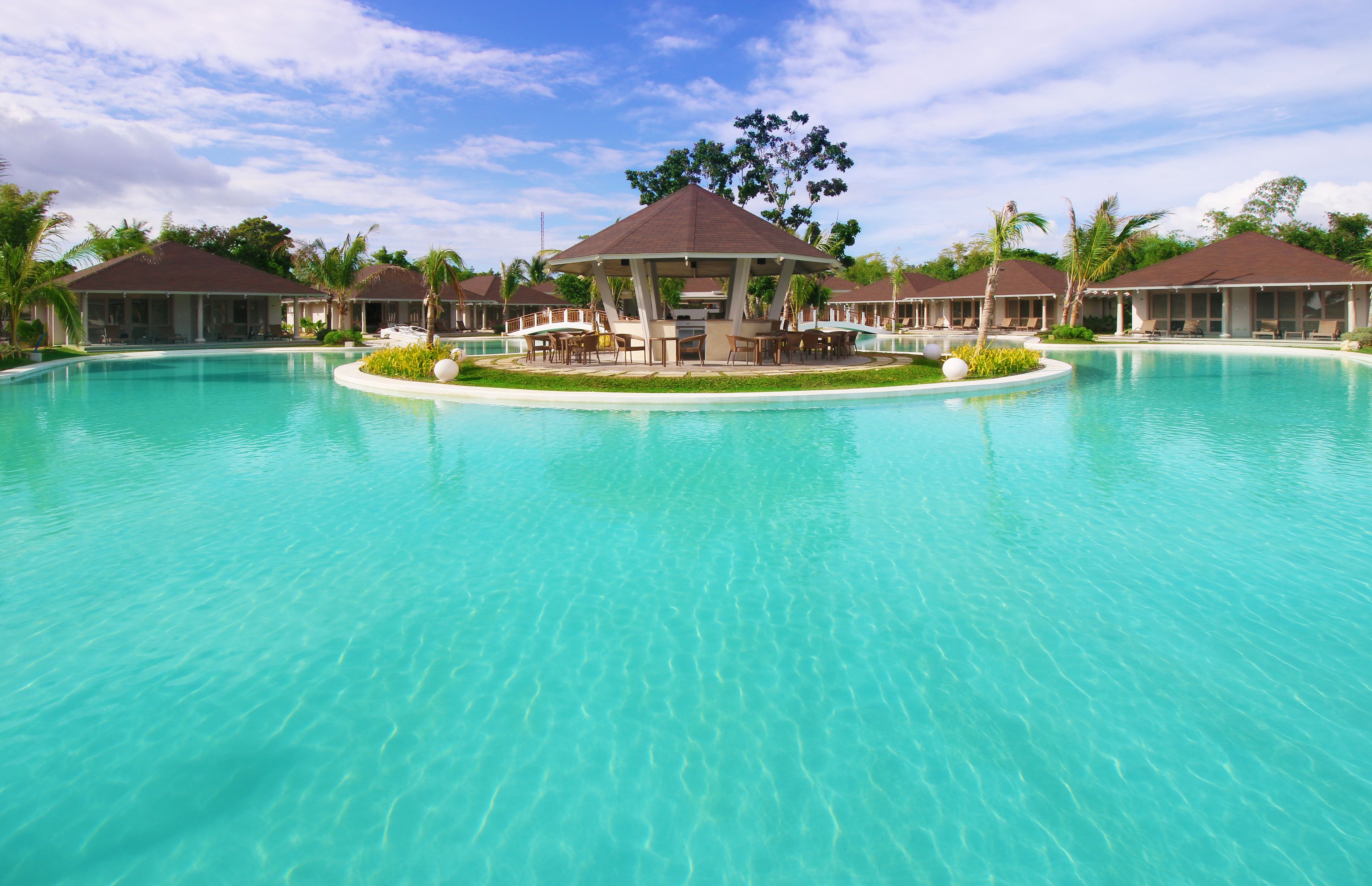 A resort pool with a central gazebo, lounge chairs, and palm trees.