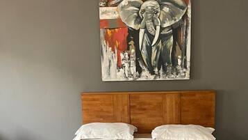 A bedroom with a wooden headboard, a blue bedspread, and a painting of an elephant on the wall.
