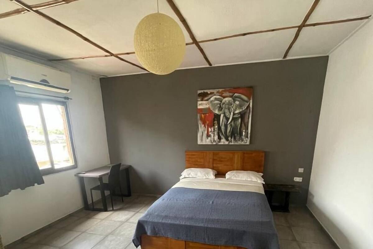 A bedroom with a bed, a desk, a chair, a hanging light fixture, and a painting on the wall.