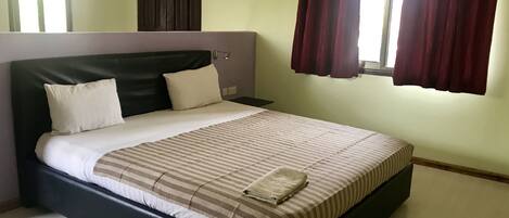 Una cama bien hecha con colcha a rayas y dos almohadas, cabecera oscura, mesita de noche y ventana con cortinas rojas.