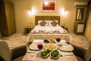A hotel room with a neatly made bed, a dining table set with fruits and a glass of red wine, and a small refrigerator.