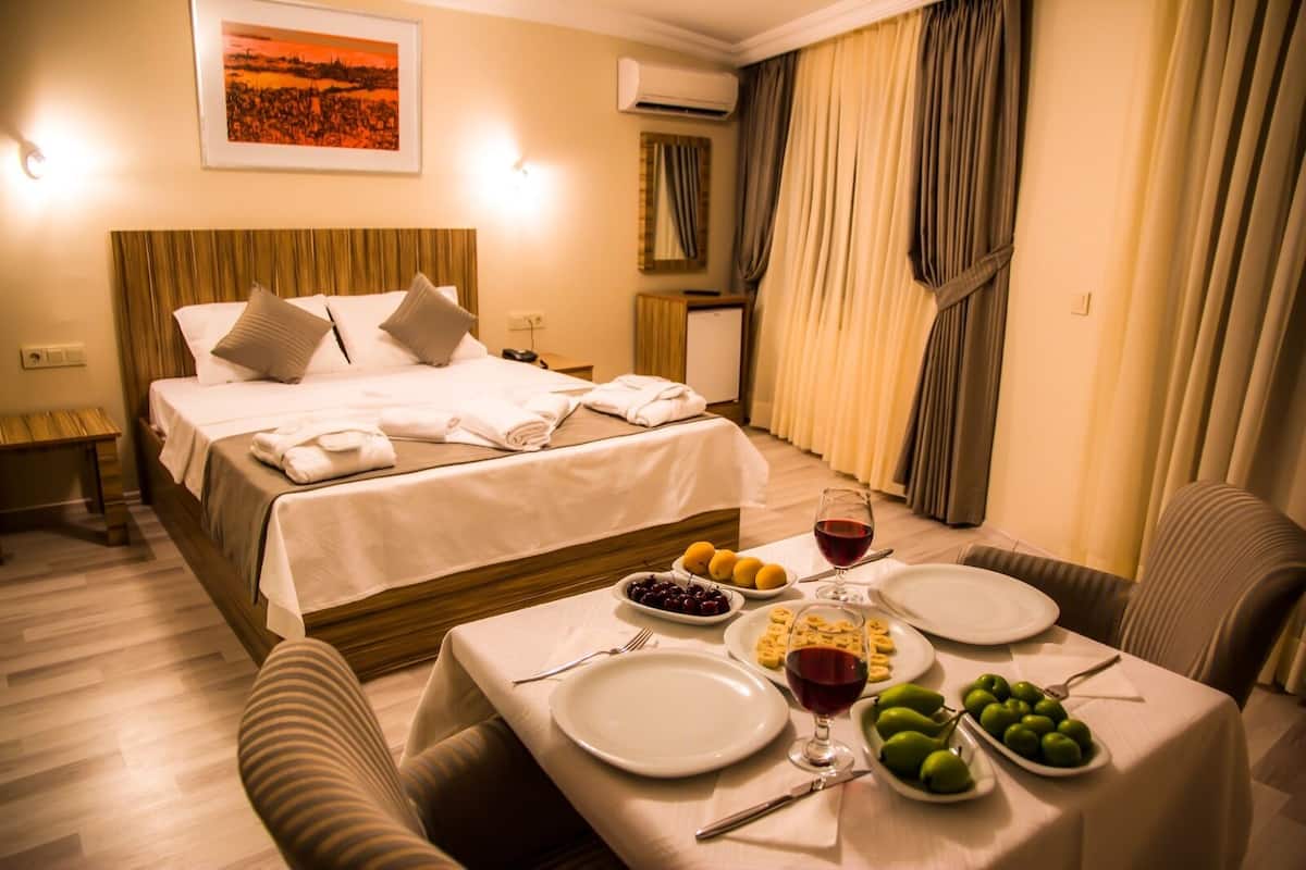 Habitación de hotel con una cama, una mesa de comedor dispuesta con platos, vasos y frutas, y un cuadro en la pared.