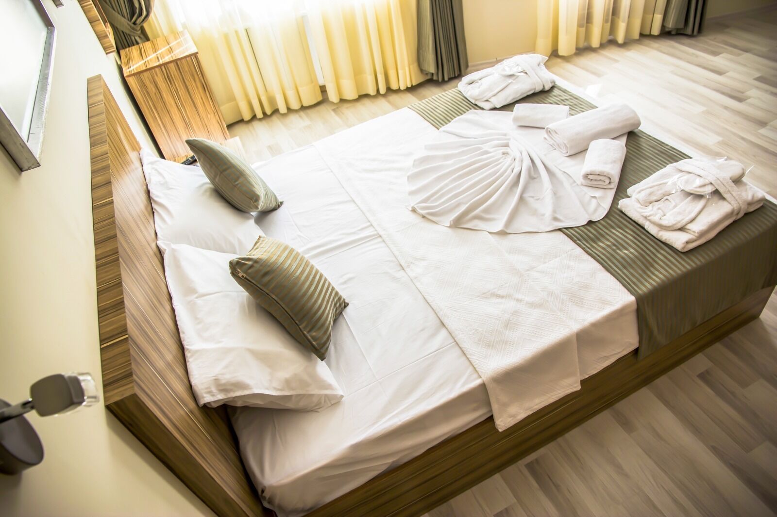 Een net opgemaakt bed met witte lakens, een groen gestreept kussen en opgerolde handdoeken op een houten bedframe in een hotelkamer.