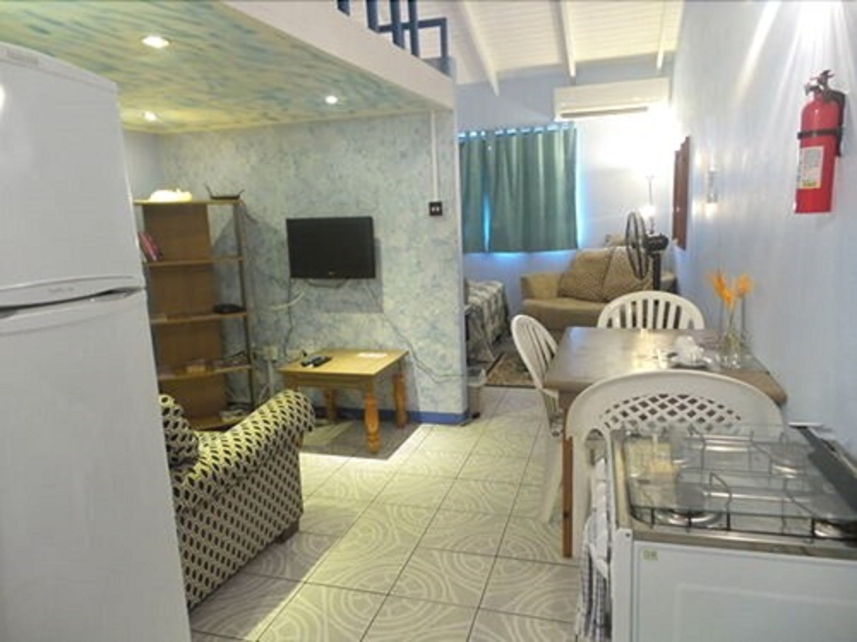 A compact kitchen with a refrigerator, a dining table with chairs, a sofa, and a television mounted on the wall.