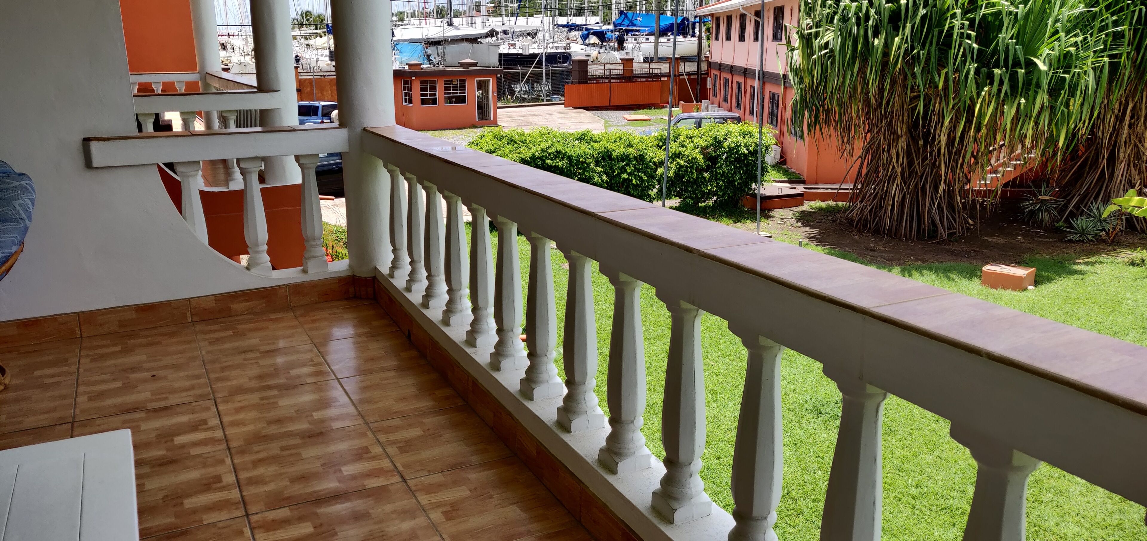Un balcón con suelo de madera y barandilla blanca, con vista a un jardín que incluye una planta grande y un edificio pequeño.