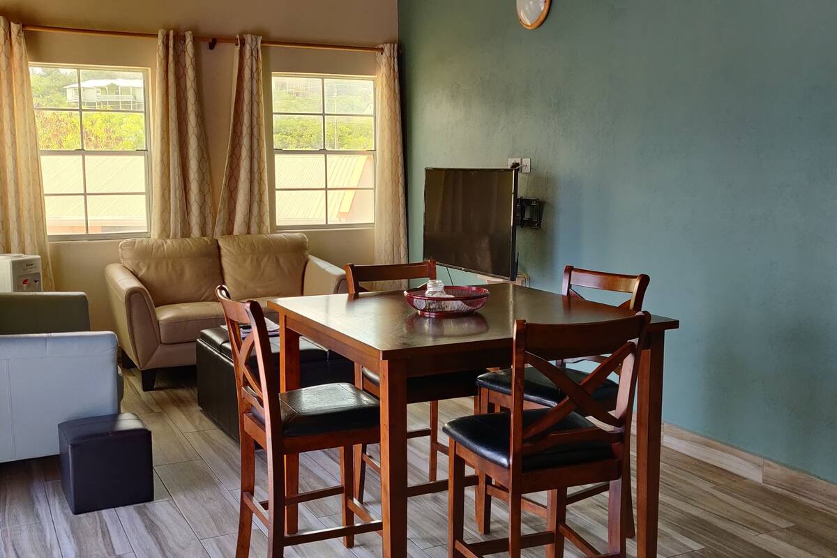 A room with a wooden dining table, four wooden chairs, a sofa, a small black ottoman, a heater, and a window with curtains.