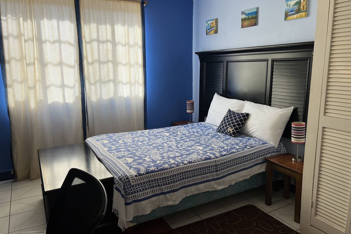A bedroom with a bed, a desk, and a chair. There are curtains, a headboard, and a nightstand with a lamp.