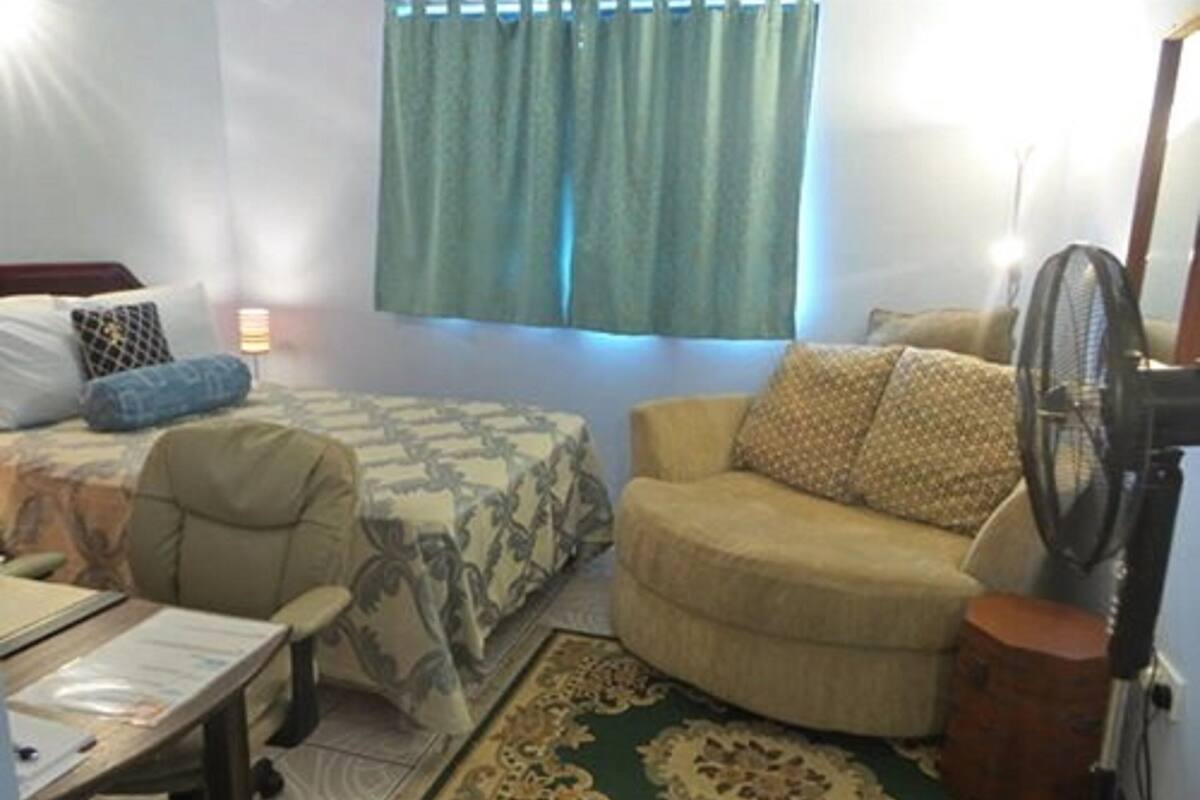 A small, compact room with a bed, a chair, a desk, and a fan.