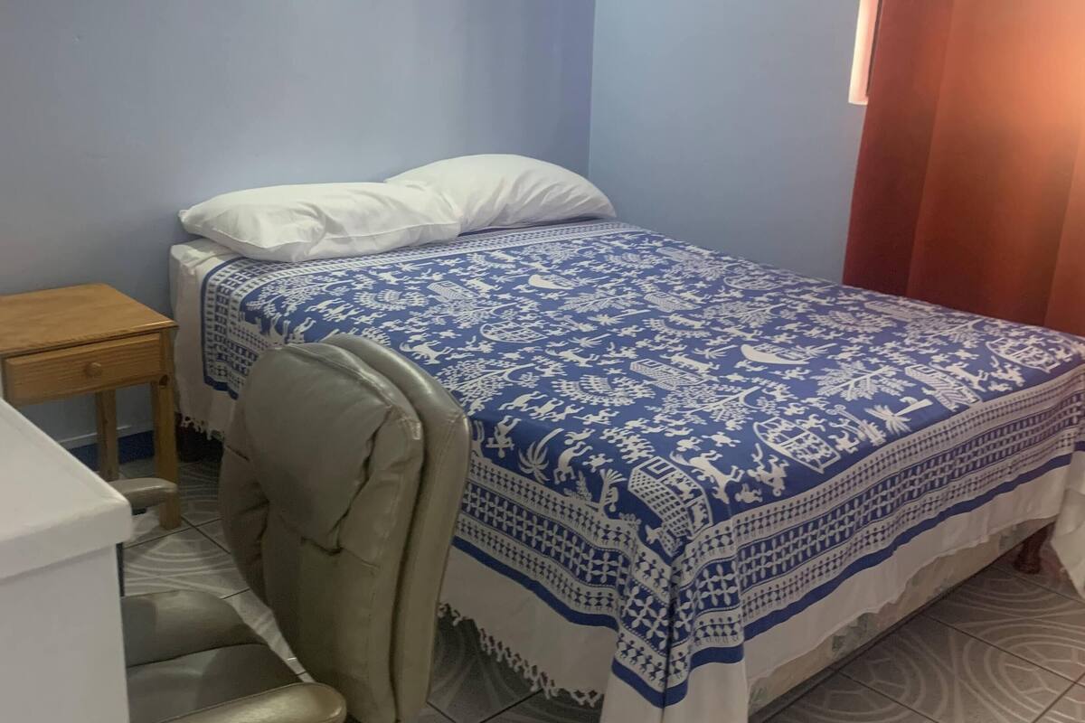 A bedroom with a bed, a chair, a small table, and a window with curtains.