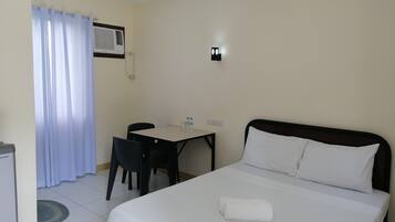 Habitación de hotel con cama, escritorio y silla. Hay una ventana con cortinas, un equipo de aire acondicionado y un teléfono en la pared.