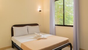 A single bed with a wooden frame, white bedding, and a white pillow. There is a window with white curtains, and a wall-mounted light fixture.