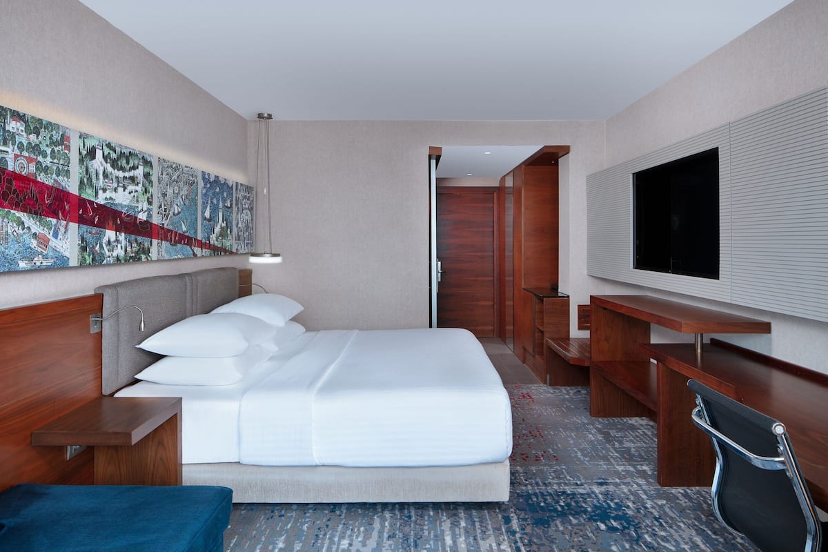A hotel room with a bed, a desk, a chair, a television, and a large abstract painting on the wall.