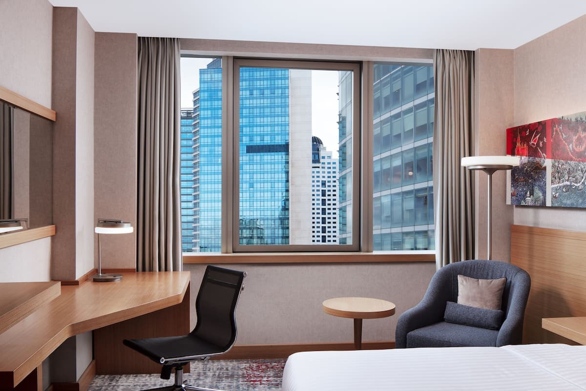 A hotel room with a large window, a desk, a chair, a bed, and a small round table.