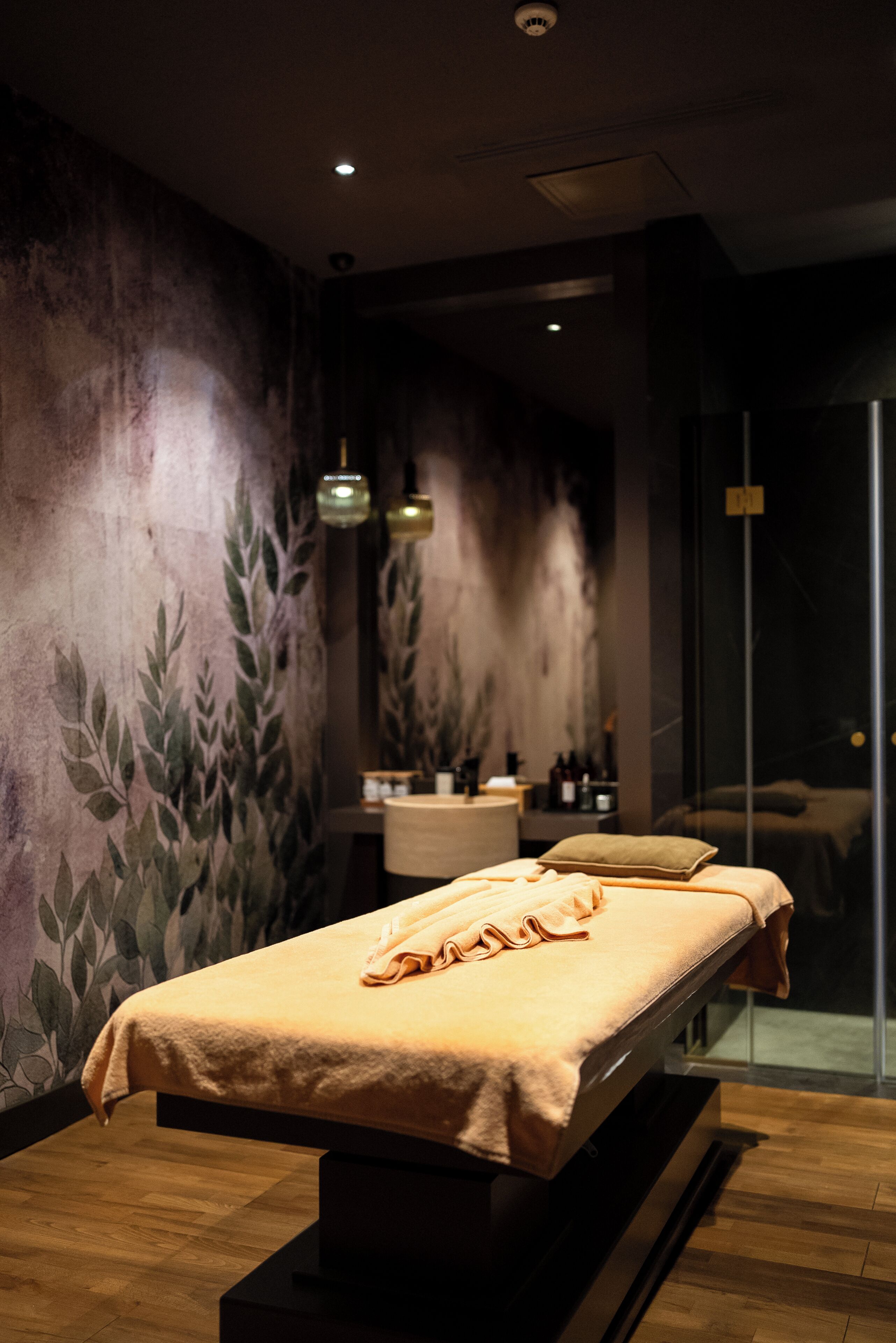 A modern spa room with a massage table, a wall with a leafy pattern, and a glass door leading to a shower area.