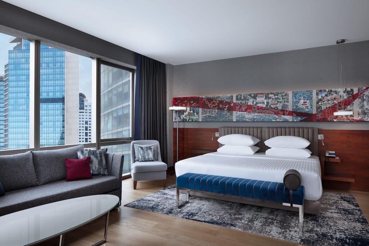 A modern hotel room with a large bed, a sofa, a chair, and a city view through large windows.