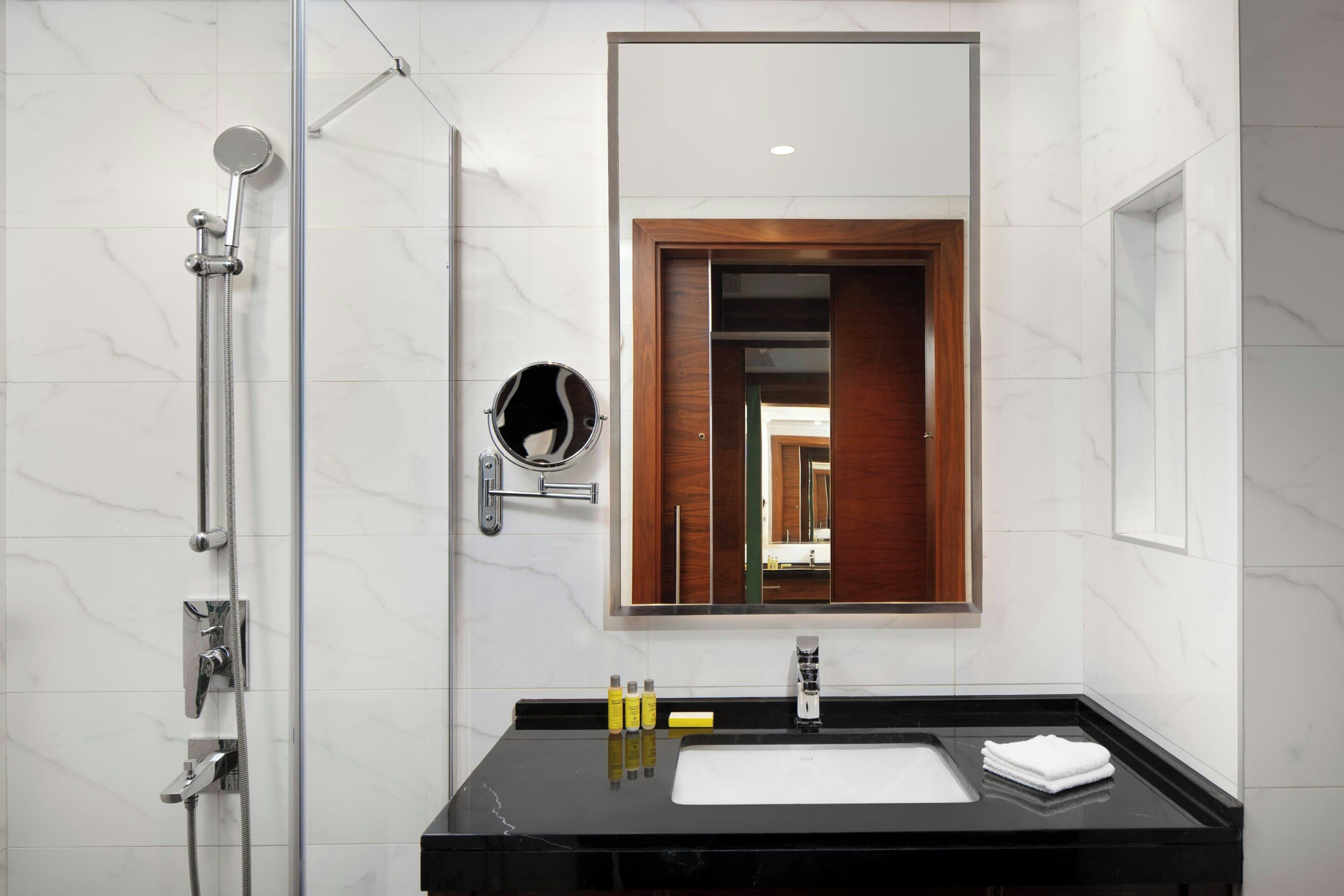 A modern bathroom with a glass-enclosed shower, a wooden-framed mirror, and a black countertop with toiletries.