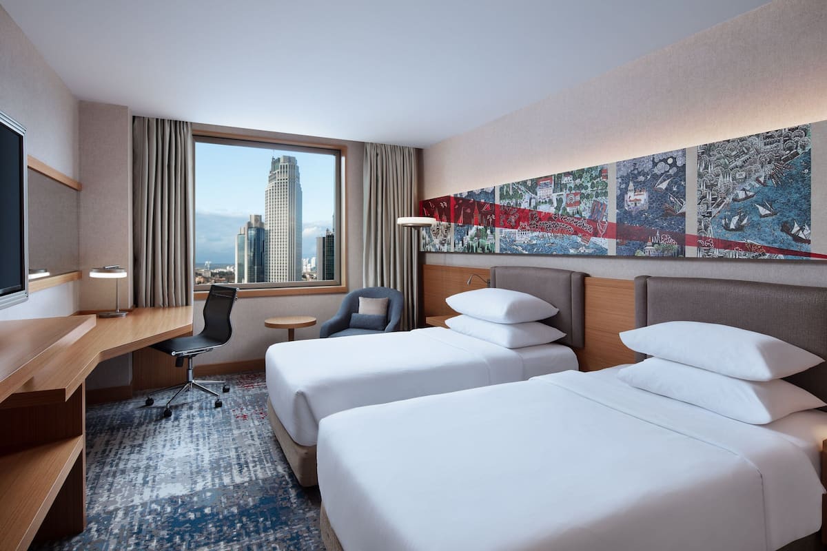 A hotel room with two beds, a desk, a chair, a TV, and a large window with a cityscape view.