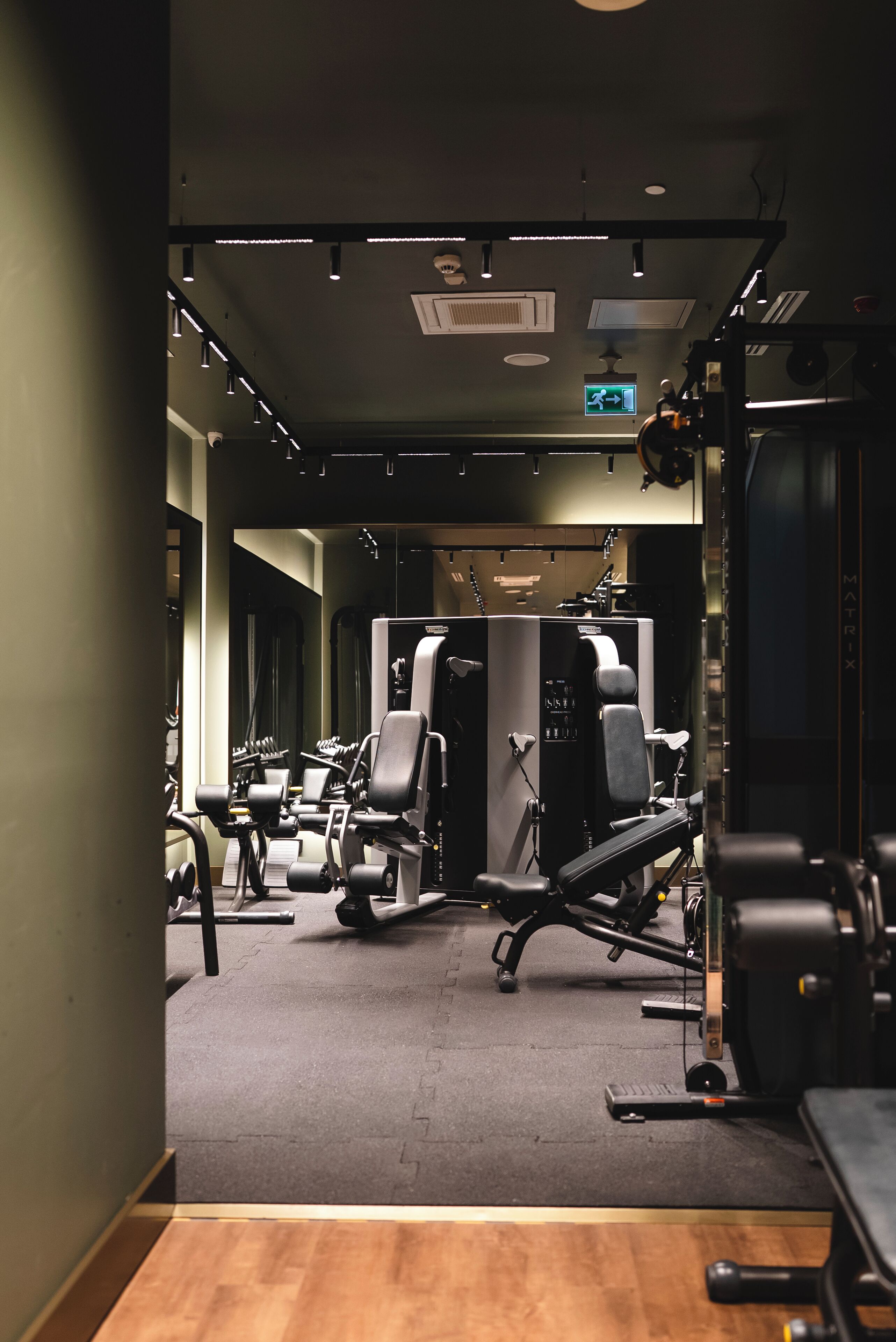 A gym with various exercise machines, a mirrored wall, and a ceiling-mounted emergency exit sign.
