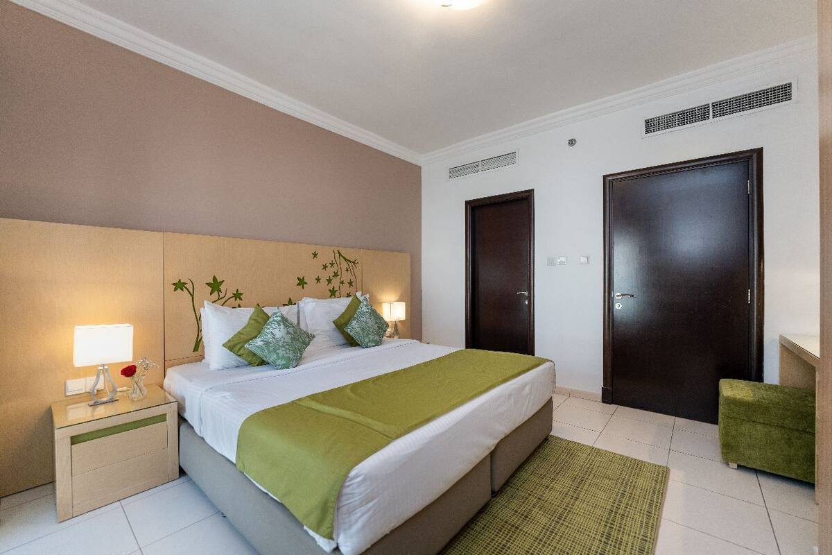 A neatly arranged hotel room with a large bed, bedside tables, a nightstand, a green throw blanket, and two doors.
