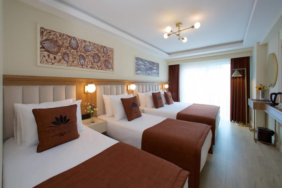 Habitación de hotel con tres camas, un cuadro grande en la pared, una lámpara en el techo y una ventana con cortinas.