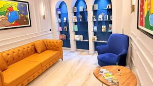 Un vestíbulo con un sofá naranja con capitoné, un sillón azul y una mesa de centro de madera con revistas. Se observan estanterías azules con libros y objetos decorativos.