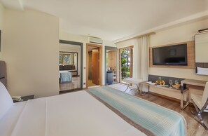 A hotel room with a large bed, a desk with a chair, a television, and a balcony with a view of greenery.