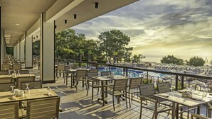 An outdoor terrace with tables and chairs, overlooking a pool and trees. - Kimeros Park Holiday Village (Kemer)