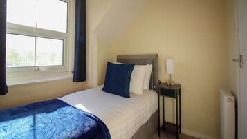 Un dormitorio con cama, mesita de noche con lámpara, ventana con cortinas y radiador.