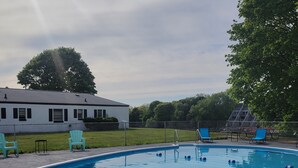 A swimming pool with blue water, surrounded by a fence. There are two blue chairs and a small table near the pool. A white building with black shutters is visible in the background. - Riverside Motel and Marina Manistee (Manistee)